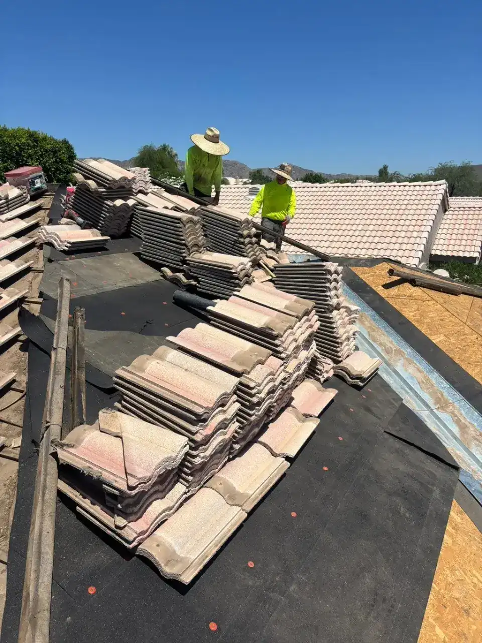 Two crew members in sun hats on a roof, with tile stacked in rows on dark underlayment